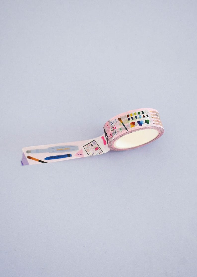 May include: A roll of light pink washi tape featuring a repeating pattern of art supplies including paintbrushes, pens, and a watercolor palette. The text "I AM AN ARTIST" is printed in a colorful font along the tape, which is partially unrolled.