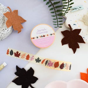 May include: A roll of washi tape labeled Sarah Frances Autumn Leaves with a pattern of colorful fall foliage. The tape features various leaf shapes in shades of red, orange, brown, and black on a cream background, accompanied by felt leaf cutouts.