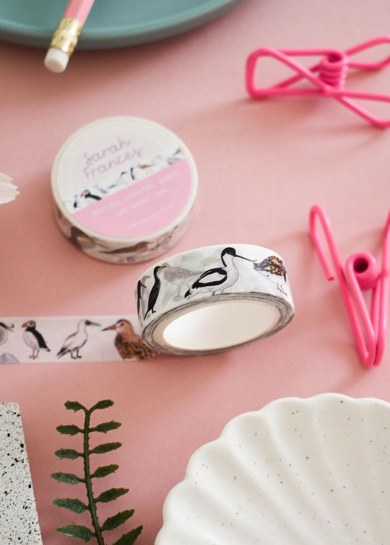 May include: A roll of decorative washi tape featuring watercolor illustrations of various coastal birds, including a puffin and an avocet. The tape is white with a repeating pattern of birds in black, white, and brown tones. The label reads Sarah Frances.