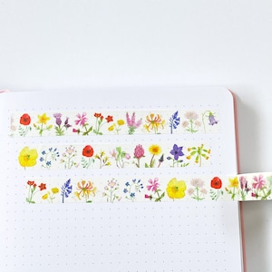 Pode incluir: Tr&ecirc;s rolos de fita washi decorativa com ilustra&ccedil;&otilde;es em aquarela de v&aacute;rias flores silvestres. A fita &eacute; branca com um padr&atilde;o repetido de flores coloridas, incluindo papoilas, narcisos e lavanda. A fita &eacute; exibida em um caderno branco com uma borda rosa.