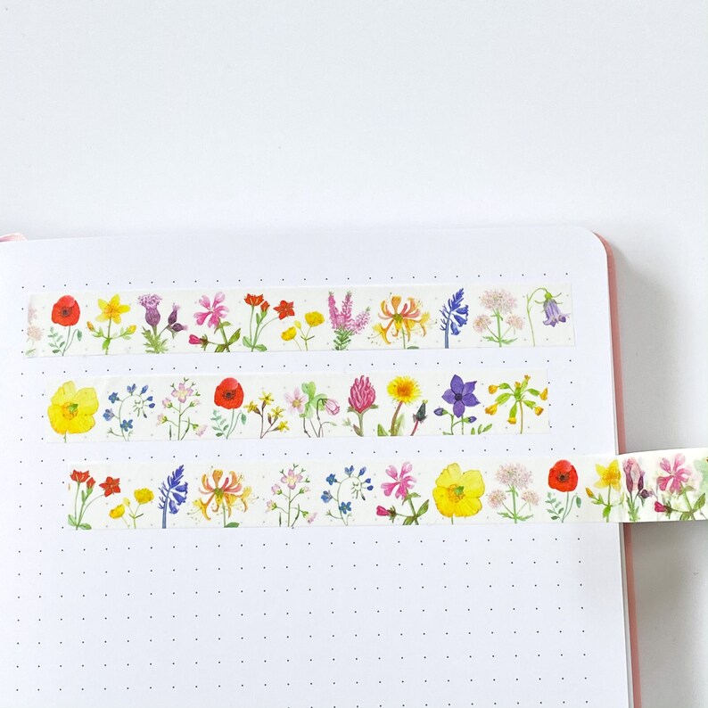 Pode incluir: Tr&ecirc;s rolos de fita washi decorativa com ilustra&ccedil;&otilde;es em aquarela de v&aacute;rias flores silvestres. A fita &eacute; branca com um padr&atilde;o repetido de flores coloridas, incluindo papoilas, narcisos e lavanda. A fita &eacute; exibida em um caderno branco com uma borda rosa.
