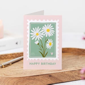 Pu&ograve; includere: Un biglietto di auguri di compleanno rosa e verde con un'illustrazione di margherita bianca e il testo "APRIL DAISY" e "HAPPY BIRTHDAY".
