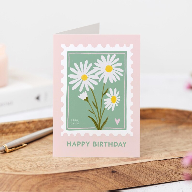 Pu&ograve; includere: Un biglietto di auguri di compleanno rosa e verde con un'illustrazione di margherita bianca e il testo "APRIL DAISY" e "HAPPY BIRTHDAY".