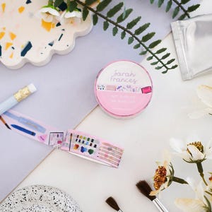 May include: A roll of pink washi tape labeled Sarah Frances Art Supplies with a pattern of watercolor paints, brushes, and pencils. The tape is shown both as a sealed roll and unrolled, displaying a repeating graphic of various art tools.
