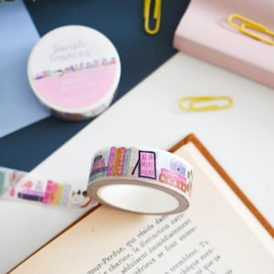 Op de afbeelding: Een rol decoratieve washi-tape met een kleurrijke illustratie van gestapelde boeken en een bordje met de tekst READ MORE BOOKS. De tape ligt op een open bladzijde van een boek, met een tweede rol en gele paperclips in de nabijheid.
