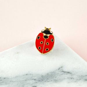 Peut inclure: Une épingle en forme de coccinelle rouge avec des points noirs et un contour doré. La tête de la coccinelle est noire, posée sur une surface en marbre blanc et gris.