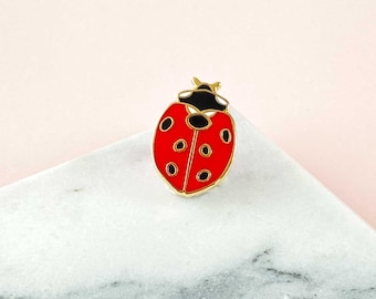 以下が含まれることがあります： 黒い斑点と金色の縁取りが付いた赤いテントウムシのピン。 テントウムシの頭は黒で、白とグレーの大理石の表面に置かれています。