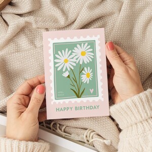 Pu&ograve; includere: Un biglietto di auguri di compleanno rosa e verde con un design di margherita bianca. Il biglietto ha un francobollo con il testo "April Daisy" e un piccolo cuore rosa. Il testo "Buon compleanno" &egrave; stampato in fondo al biglietto.