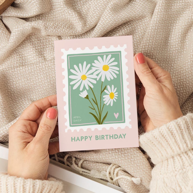 Pu&ograve; includere: Un biglietto di auguri di compleanno rosa e verde con un design di margherita bianca. Il biglietto ha un francobollo con il testo "April Daisy" e un piccolo cuore rosa. Il testo "Buon compleanno" &egrave; stampato in fondo al biglietto.
