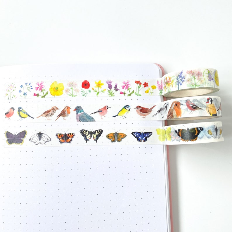 Pode incluir: Rolos de fita washi decorativa com ilustra&ccedil;&otilde;es em aquarela de p&aacute;ssaros, borboletas e flores. Os desenhos da fita incluem p&aacute;ssaros coloridos, borboletas com padr&otilde;es de asas detalhados e padr&otilde;es florais. A fita &eacute; mostrada em um caderno rosa.