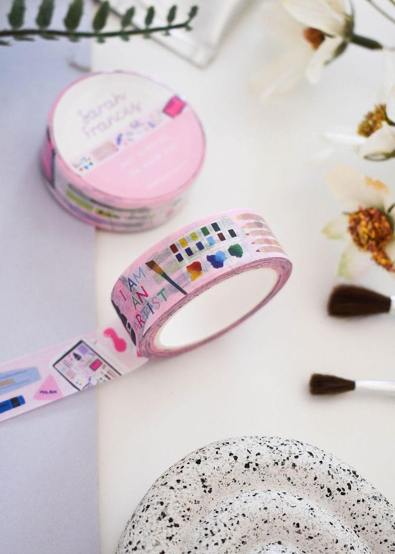 May include: Two rolls of light pink washi tape featuring artistic illustrations of paintbrushes, watercolor palettes, and colored pencils. The tape displays the text I AM AN ARTIST in colorful letters, along with various art supply graphics.
