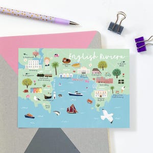 May include: A colorful illustrated map of the English Riviera, featuring landmarks, boats, and the text "English Riviera." The map is on a postcard with a pink envelope, a purple pen, and binder clips.