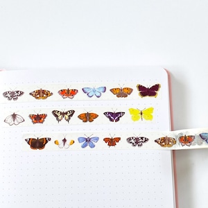 Pode incluir: Fita washi decorativa com desenhos de borboletas em tons de laranja, amarelo, azul, roxo e preto. A fita &eacute; exibida em um caderno rosa com um fundo de papel branco pontilhado. Ideal para artesanato e journaling.