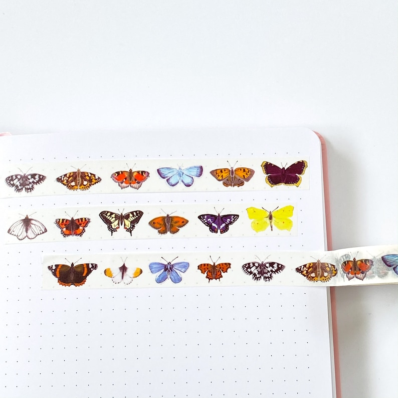 Pode incluir: Fita washi decorativa com desenhos de borboletas em tons de laranja, amarelo, azul, roxo e preto. A fita &eacute; exibida em um caderno rosa com um fundo de papel branco pontilhado. Ideal para artesanato e journaling.