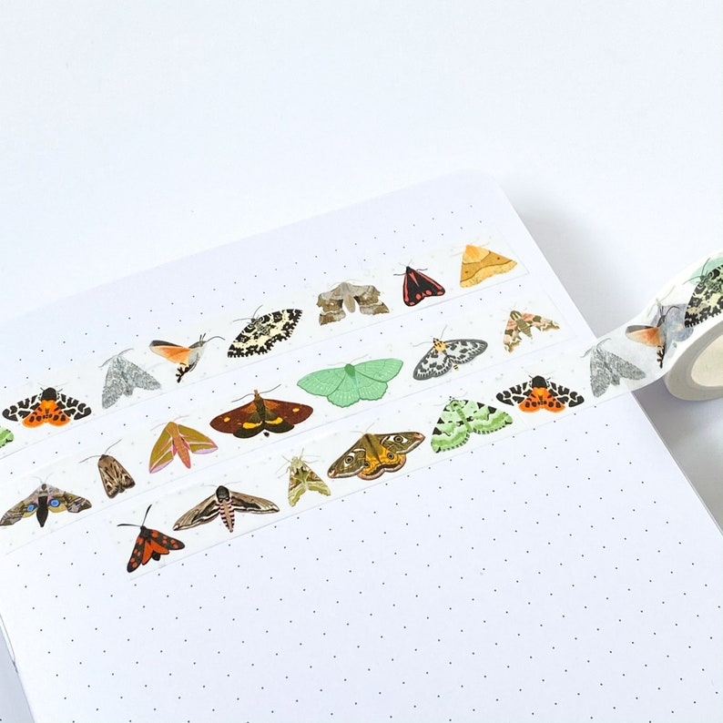 May include: A roll of decorative adhesive tape featuring a repeating pattern of various moth species in diverse colors including orange, green, brown, and black, applied to a white dotted journal page with a portion of the roll visible on the right.