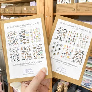 May include: Two boxes of British Nature Greetings Cards, Volume One and Two, are held. Each box is brown with a white label featuring illustrations of British wildlife. The text on the boxes reads "Includes 6 cards and kraft envelopes" and "Designed and printed in the UK."