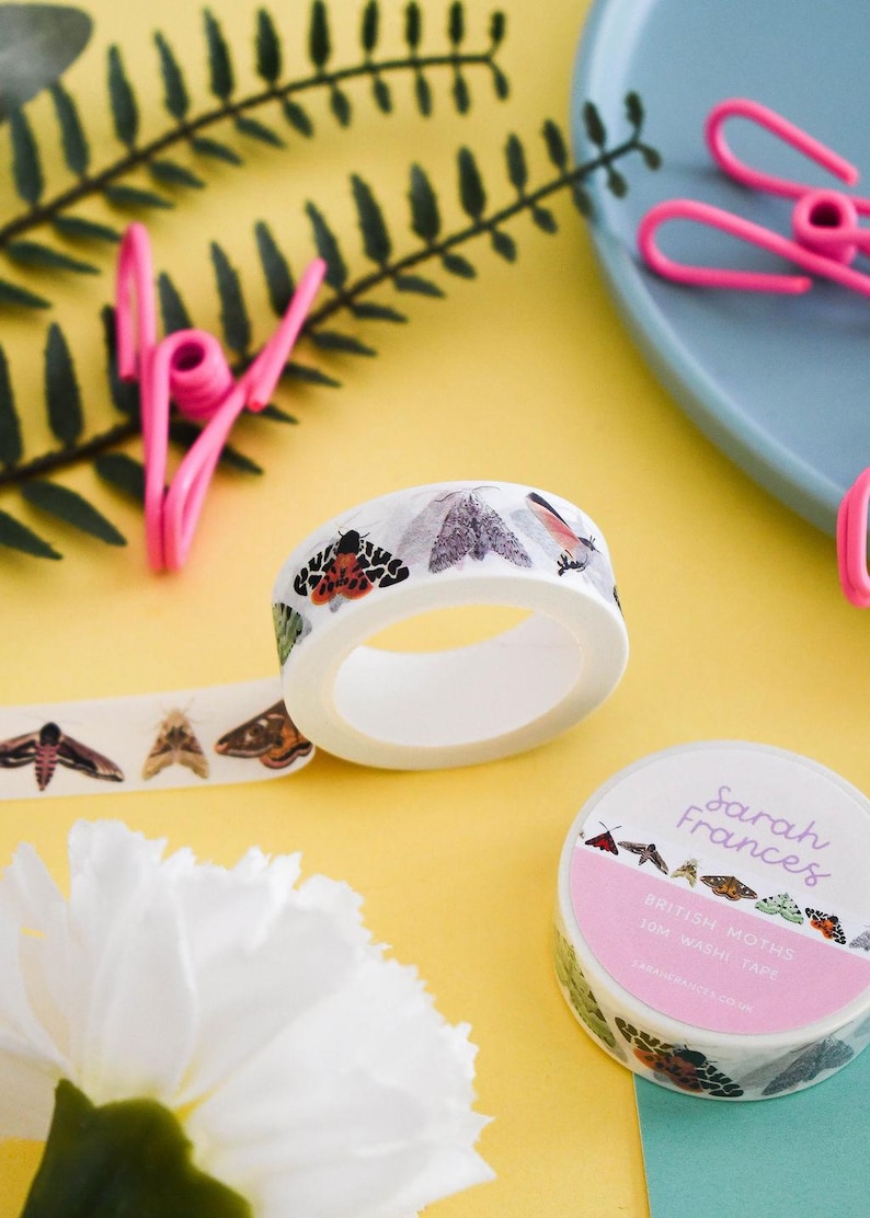 May include: A roll of white washi tape featuring a repeating pattern of various colorful moths. The label on the roll reads Sarah Frances, British Moths, 10m Washi Tape. The tape is shown both as a roll and partially unrolled to display the moth illustrations.