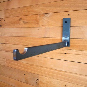 May include: A dark gray metal shelf bracket mounted on a wooden wall. The bracket has a rectangular mounting plate with two holes and a horizontal arm with a curved end. The wood has a natural grain.