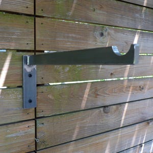 May include: A metal shelf bracket mounted to a wooden wall. The bracket is made of a single piece of metal that is bent to form a shelf support and a mounting plate. The bracket is painted a dark gray color.