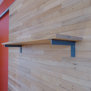 May include: A wooden shelf with a light brown finish is mounted on a wall made of light brown wooden planks. The shelf is supported by two blue metal brackets. The wall is adjacent to an orange wall.