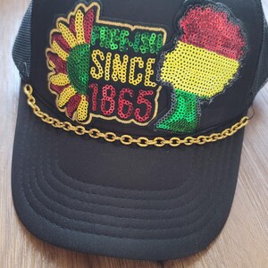May include: A black trucker hat featuring a sequined patch with a sunflower and a silhouette of the African continent in red, yellow, and green. The text reads "FREE-ISH SINCE 1865" in yellow. A gold-colored chain trim sits across the brim base.