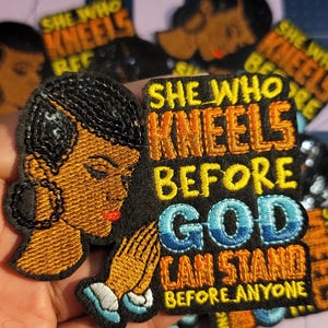 May include: Embroidered patch featuring a woman's profile and the text "SHE WHO KNEELS BEFORE GOD CAN STAND BEFORE ANYONE." The patch has black, orange, and blue stitching. The woman's profile is in profile with a red lip.
