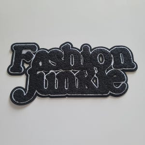 May include: A black and white patch with the words "Fashion Junkie" in a bold, stylized font. The letters are black with a textured appearance and a white outline, creating a striking contrast. The patch is likely for clothing or accessories.