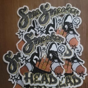 May include: A collection of embroidered patches with a sneaker theme. The patches feature the word "Sneaker" in a stylized font, along with images of sneakers and the text "Head Lad". The patches are in shades of black, white, orange, and gold.