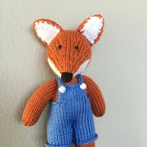 May include: A knitted fox toy wearing blue overalls. The fox has orange fur, a black nose, and white ears.