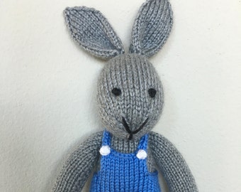 May include: A gray knitted bunny wearing blue overalls with white buttons. The bunny has black eyes and a black nose.
