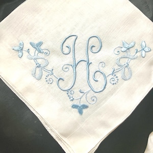 May include: A white square handkerchief featuring a central embroidered letter H in light blue thread. The monogram is flanked by symmetrical light blue floral and vine embroidery patterns that extend outward across the fabric surface.