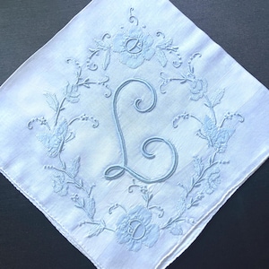 May include: White handkerchief with a light blue embroidered floral design and the letter "L" in the center. The embroidery features flowers and leaves, creating a delicate, vintage aesthetic.