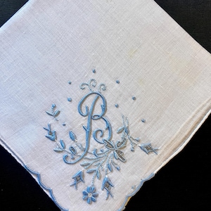 May include: A square white handkerchief featuring a light blue embroidered letter B in the center, surrounded by delicate floral and leaf motifs. The edges of the fabric are finished with a scalloped light blue stitched border detail.