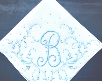 May include: A square white handkerchief featuring light blue embroidery. The center displays a large, ornate letter B in cursive script, surrounded by delicate floral vines, leaves, and small dots that extend toward the scalloped edges.