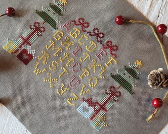 May include: A cross-stitch pattern on a gray fabric background with a Christmas theme. The pattern features a circle of letters spelling out "Merry Christmas" with three stylized Christmas trees and presents.