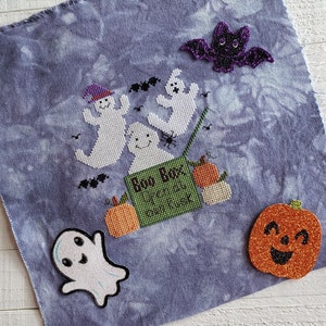 May include: A blue fabric with a Halloween-themed cross-stitch design. The design features a green box with the text "Boo Box Treats & Tricks", three white ghosts, a black bat, and two orange pumpkins.