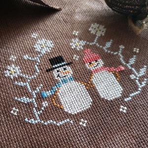May include: Cross-stitch embroidery of two snowmen holding hands, standing in front of a brown background with white flowers and blue branches.