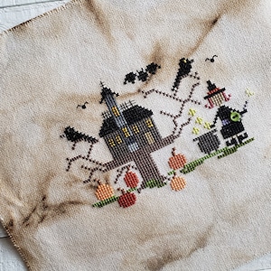 Op de afbeelding: Kruissteekontwerp met een Halloween-scène. Een zwart spookhuis met gele ramen staat naast een boom met vleermuizen en kraaien. Een heks roert in een ketel, met pompoenen aan de voet van de boom. Het ontwerp staat op een bruinachtige stof.