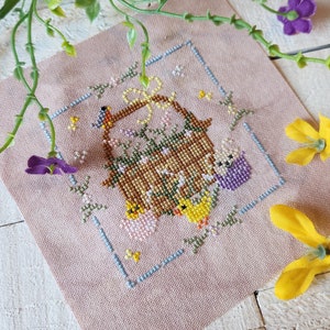 May include: A cross-stitch embroidery of a basket filled with chicks and flowers. The basket is brown with a white bow and is surrounded by a blue frame. The background is pink.
