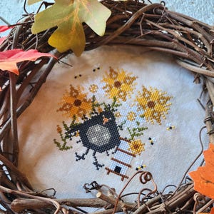Könnte beinhalten: Ein Kreuzstich-Design mit einem schwarzen Vogel mit einem Sonnenblumenstrauß und einem kleinen Bienenstock. Das Design befindet sich in einem natürlichen Weinrebenkranz, der mit bunten Herbstblättern geschmückt ist. Die Stickerei befindet sich auf cremefarbenem Stoff.