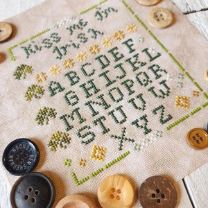 Puede incluir: Un sampler de punto de cruz con el texto "Kiss me I'm Irish" y el alfabeto en letras verdes. El sampler está rodeado de botones de madera de varios colores y tamaños.