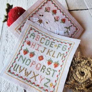 May include: Two cross-stitch embroidery patterns on white fabric. The top pattern features a pink background with strawberries, bees, and a white bird. The bottom pattern features a white background with the alphabet in green and orange letters, surrounded by strawberries.