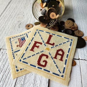 May include: Two cross-stitched fabric squares, one with an American flag and the other with the letters "FLAG" in red, white, and blue.