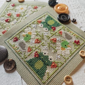 Może przedstawiać: Wzór haftu krzyżykowego w kolorze zielonym, białym i czerwonym z motywem kwiatowym. Wzór jest oprawiony w zieloną ramkę i otoczony guzikami.