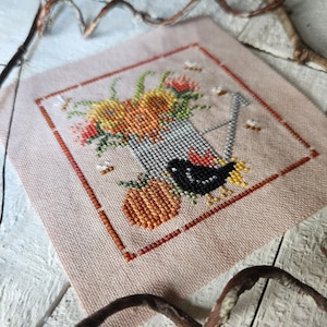 Könnte beinhalten: Ein Kreuzstichmuster mit einem schwarzen Vogel, der in einem Eimer mit orangefarbenen, gelben und roten Blumen sitzt. Der Eimer ist von einem roten Rahmen umgeben.