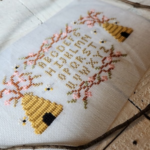 May include: Cross-stitch embroidery on white fabric with the text "Honey is sweetest in the spring" surrounded by a floral border with pink blossoms and bees.
