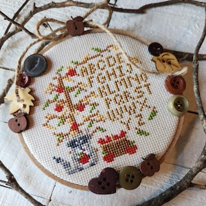 May include: A cross-stitch embroidery hoop featuring an alphabet tree design with red apples. The embroidery is surrounded by decorative wooden buttons and twine, with twigs in the background. The text "ABCDEFGHIJKLMN OPQRSTUVWXYZ" is stitched in gold thread.
