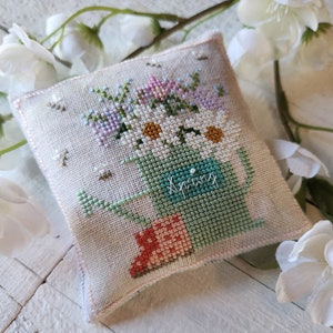 May include: A white fabric pillow with a cross-stitch design of a green watering can filled with pink, white, and purple flowers. The watering can has the word "Spring" stitched on it.