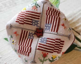 Puede incluir: Un cojín para alfileres de forma cuadrada con un fondo blanco y cuatro banderas americanas bordadas en rojo, blanco y azul. El cojín está decorado con pequeñas flores y un botón rojo en forma de estrella en el centro.