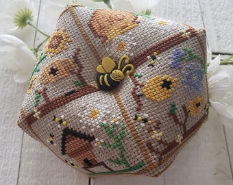 Puede incluir: Un protector de agujas hexagonal con un diseño de abeja. El protector de agujas está hecho de tela y tiene un patrón de punto de cruz de abejas, flores y panales. La abeja es amarilla y negra y tiene una cara sonriente.